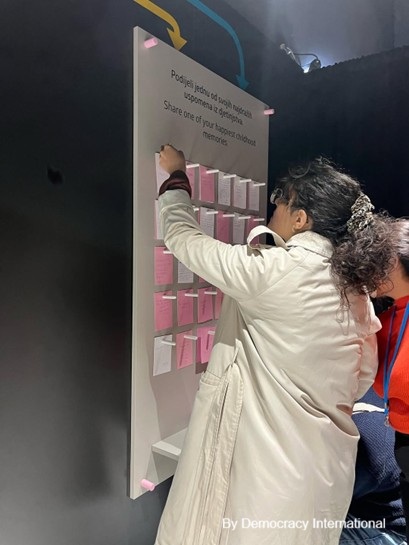 The image displays a person putting a memory written on a note on a board collecting all the memories. The header of the board is “Share one of your happiest childhood memories”.