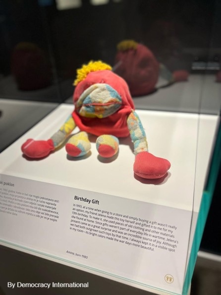 The image displays a plush with the following label: “In 1993, at a time when going to a store and simply buying a gift wasn’t really an option, my friend Jelena made this toy herself and gifted it to me for my 13th birthday. To make it, she used pieces of old clothing and other materials she found at home. Since gifts weren’t part of everyday life in wartime, Jelena’s present came as a great surprise and was an incredible source of joy. Although we had both outgrown such toys by that time, I always kept it