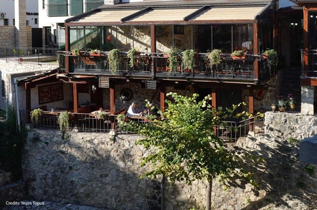 A traditional stone-built restaurant in Mostar, featuring upper and lower terraces decorated with hanging plants and morning sunlight.