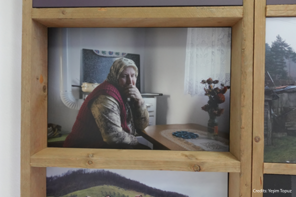 Photograph displayed at the Srebrenica museum, showing an elderly woman seated at her kitchen table in a moment of quiet reflection.