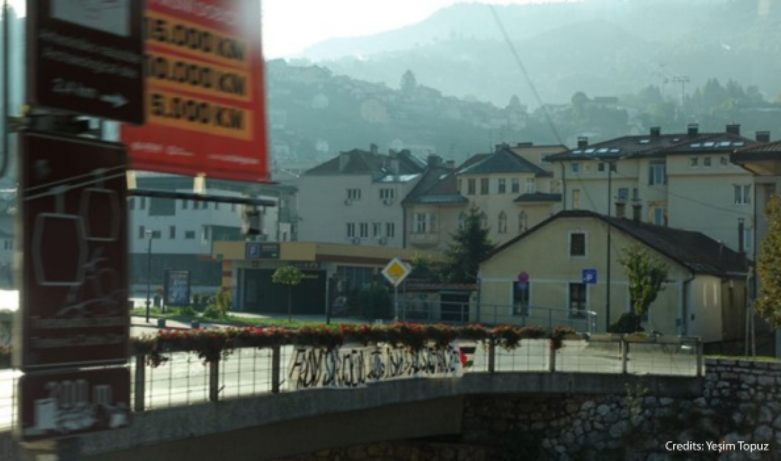 View from a moving vehicle crossing a bridge in Sarajevo, overlooking residential buildings and the fog-covered hills that surround the city.